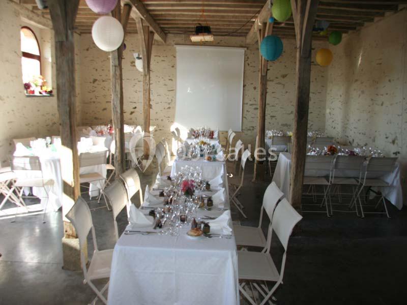 Salle rustique décorée pour un repas, avec tables dressées, chaises blanches et lanternes suspendues colorées.