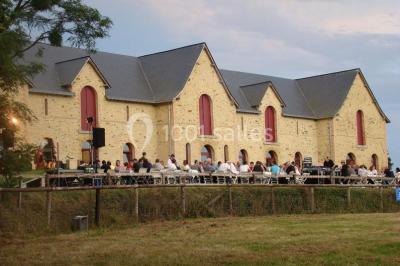 Salle de réception rustique avec tables rondes et rectangulaires dressées pour un événement, éclairage tamisé.