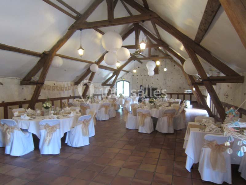 Salle de réception avec poutres apparentes, tables rondes décorées de nappes blanches et chaises ornées de nœuds beiges.