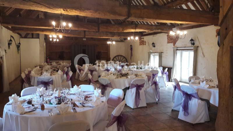 Salle de réception rustique avec poutres apparentes, tables décorées pour un événement et chaises ornées de nœuds violets.