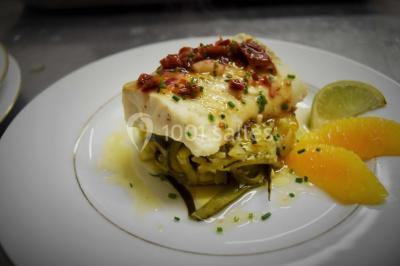 Filet de poisson servi sur un lit de légumes, garni de tomates séchées, herbes fraîches, et accompagné d'agrumes.