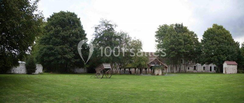 Cour verdoyante avec pelouse, arbres, charrette en bois et bâtiments anciens en pierre en arrière-plan.