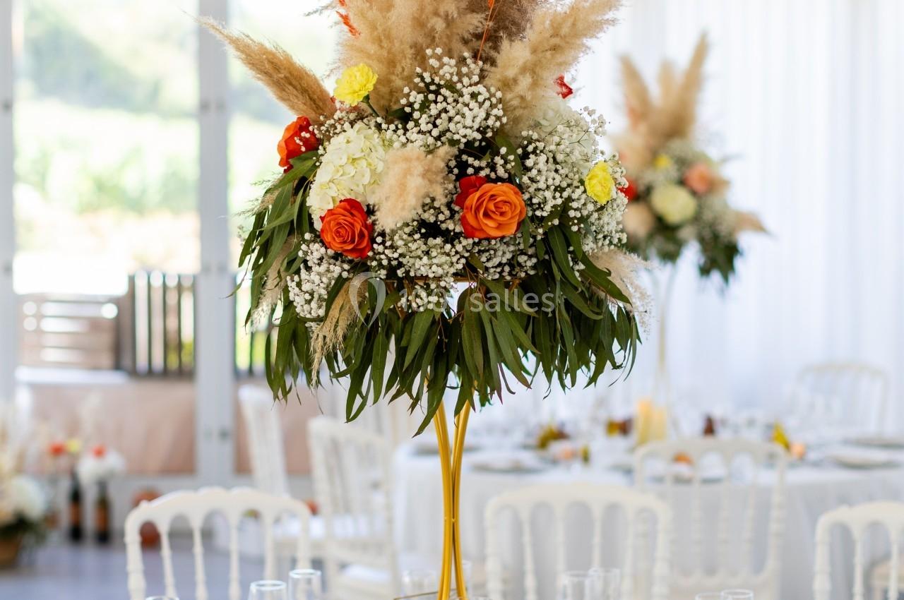 Centre de table floral avec pampas, roses orange et blanches, sur une table dressée dans une salle lumineuse.
