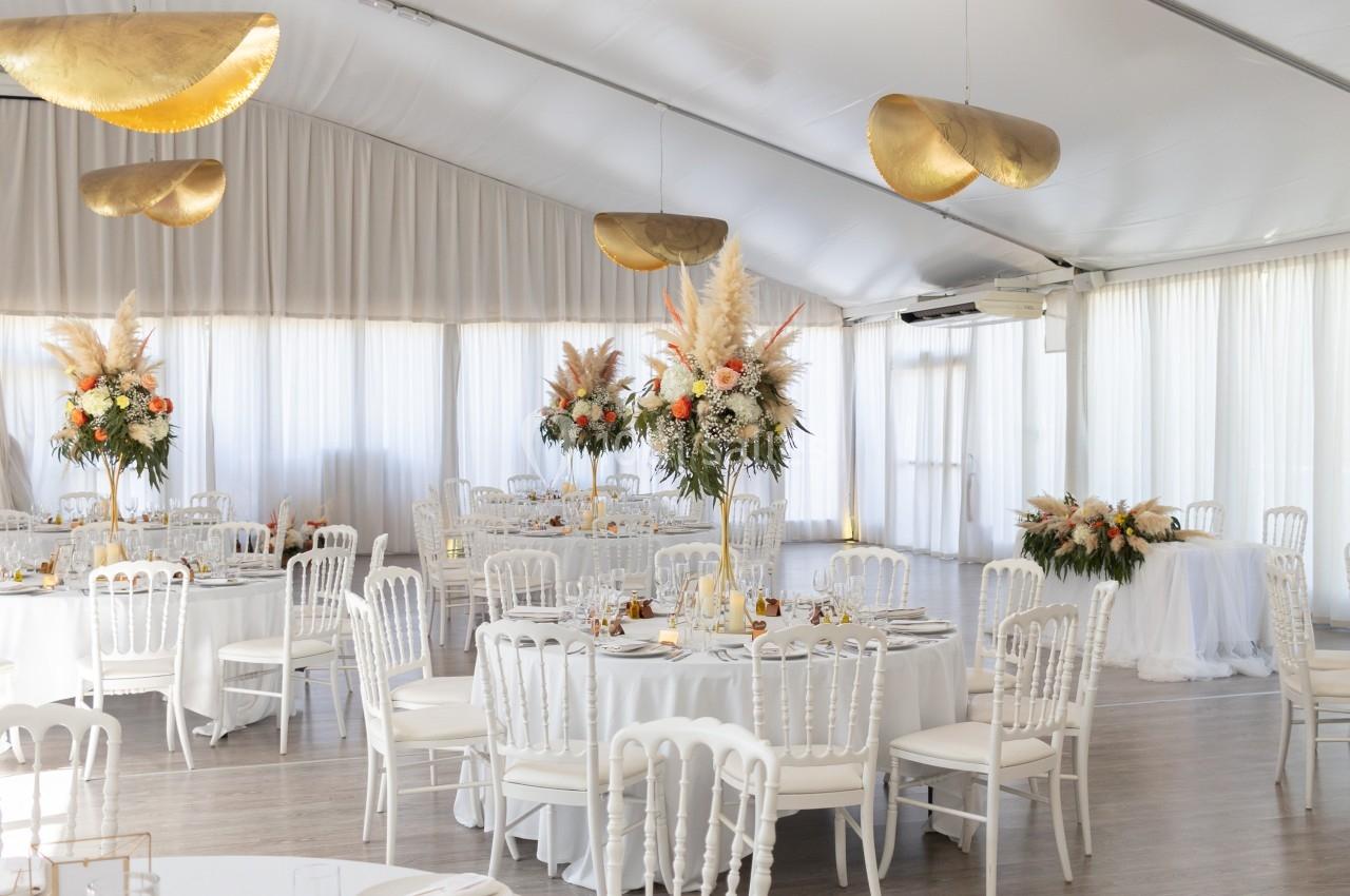Salle de réception lumineuse décorée de tables rondes avec nappes blanches, chaises élégantes et compositions florales.
