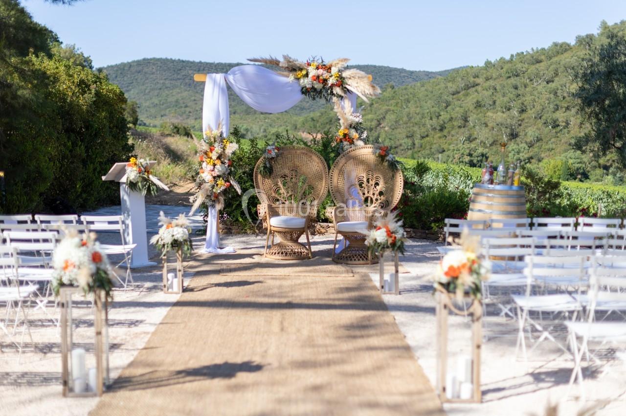 Allée décorée menant à une arche fleurie avec deux fauteuils en osier, dans un cadre naturel et verdoyant.