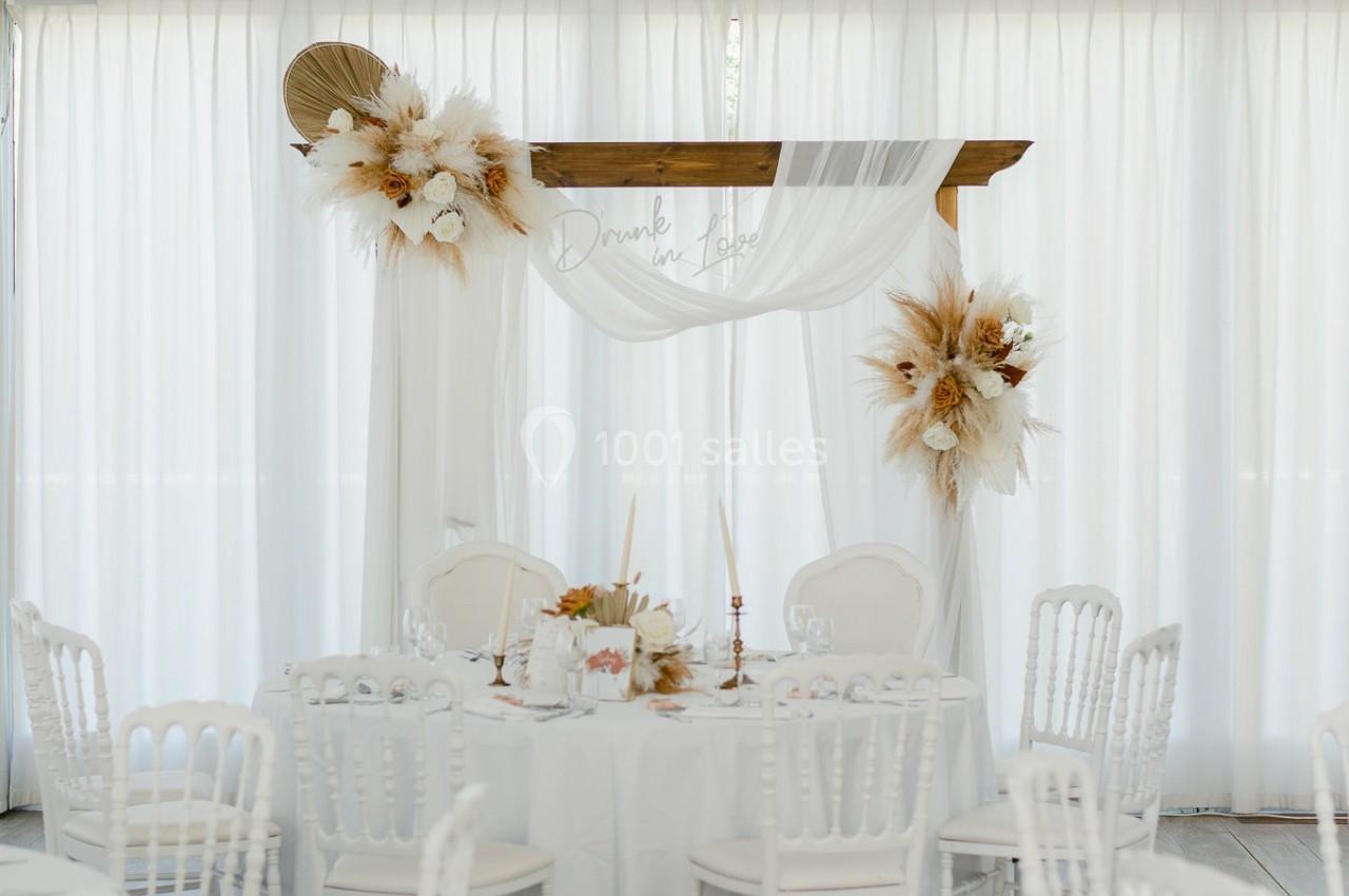 Table de réception décorée de fleurs séchées et voilage blanc, entourée de chaises blanches dans une salle lumineuse.