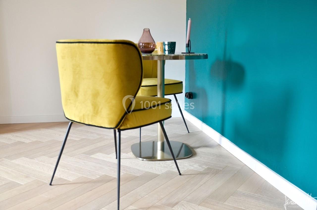 Deux chaises jaunes autour d'une table ronde en métal, posées sur un parquet clair près d'un mur bleu.