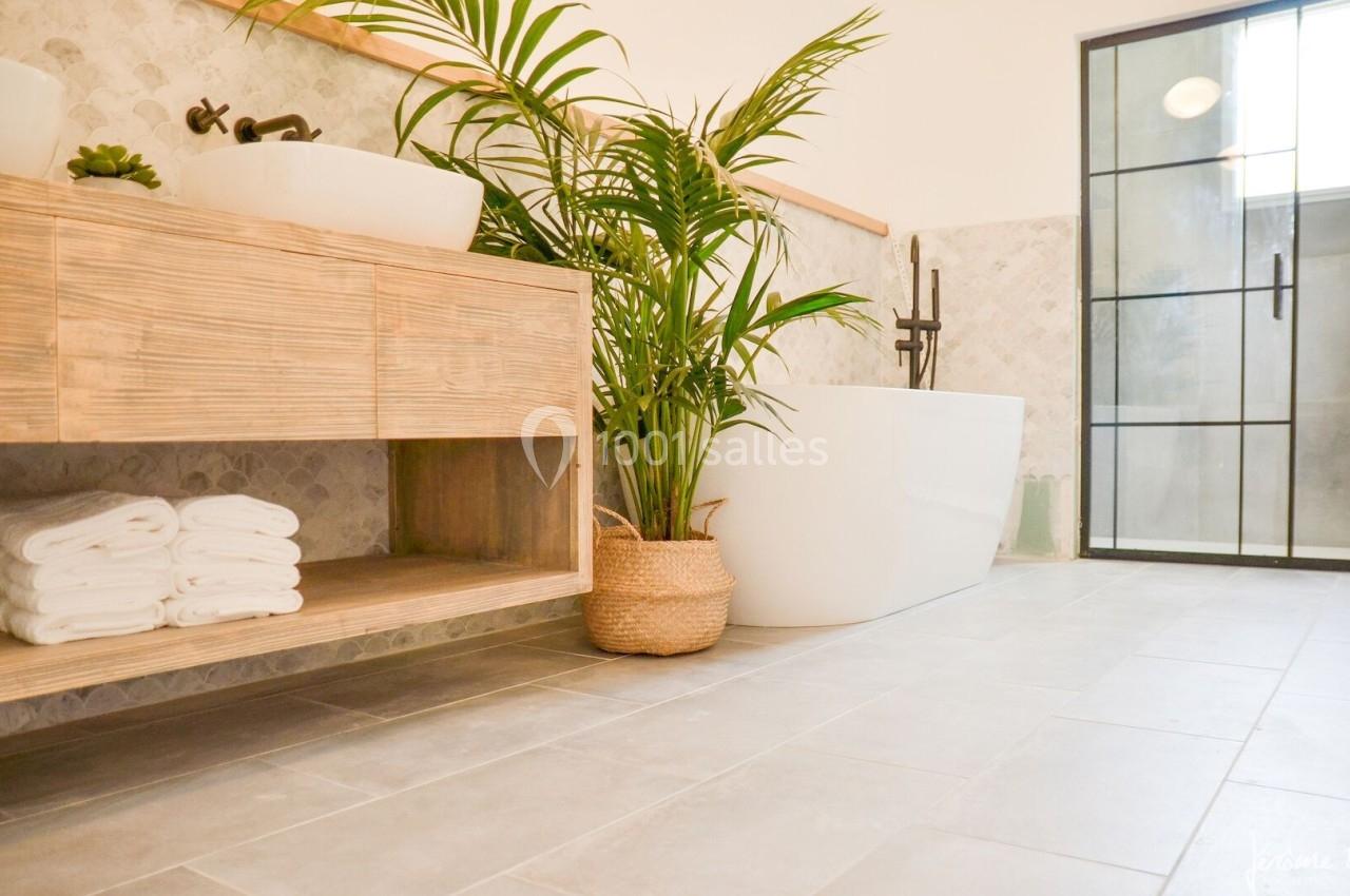 Salle de bain lumineuse avec baignoire blanche, meuble en bois, plantes et douche vitrée en arrière-plan.