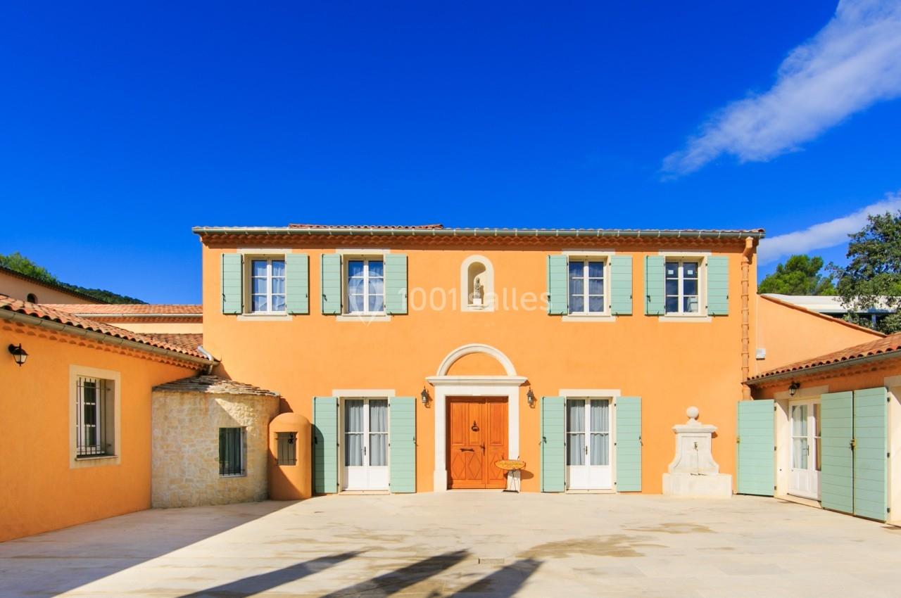 Façade d'une maison méditerranéenne avec murs orange, volets verts et cour ensoleillée sous un ciel bleu.