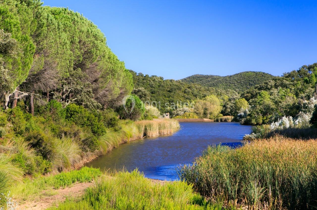 Paysage naturel avec une rivière bordée de végétation dense, entourée de collines boisées sous un ciel bleu clair.