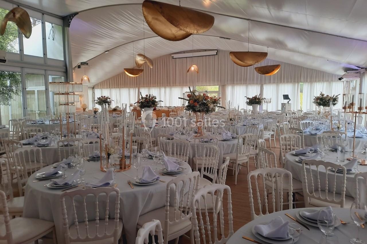 Salle de réception décorée avec des tables rondes dressées, nappes blanches, chaises élégantes et luminaires dorés.
