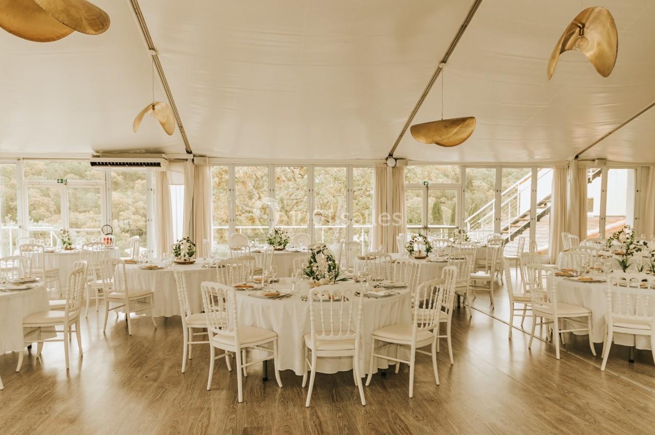 Salle de réception lumineuse avec tables rondes dressées, nappes blanches et décorations florales.