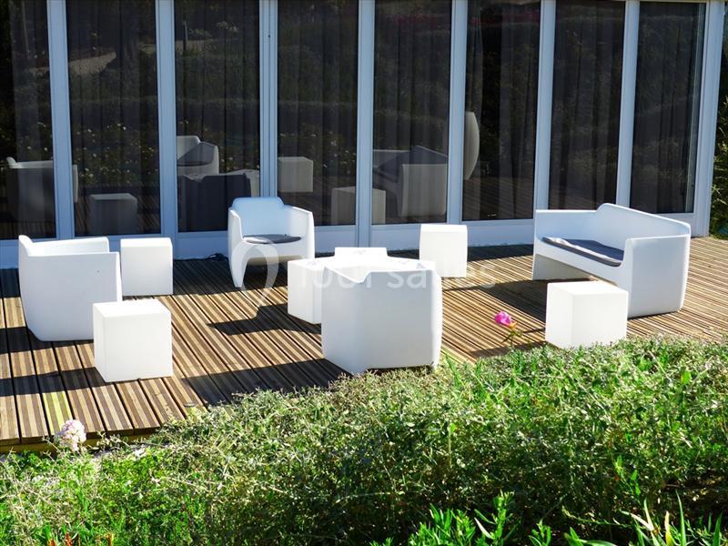 Mobilier de jardin blanc moderne disposé sur une terrasse en bois devant une baie vitrée, entourée de verdure.