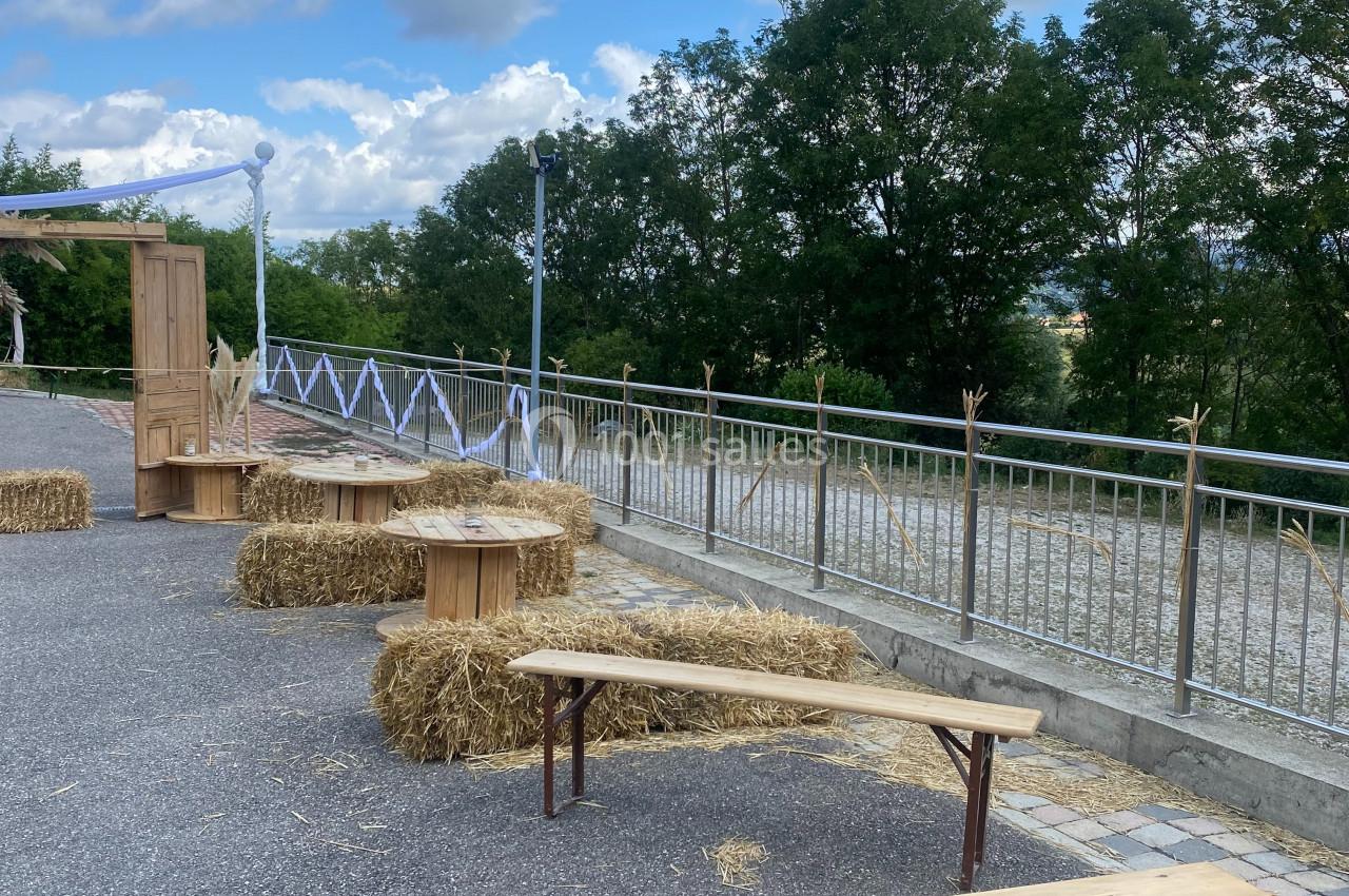 Bancs en bois et bottes de paille disposés en extérieur sur un sol pavé, avec des arbres et un ciel nuageux en arrière-plan.