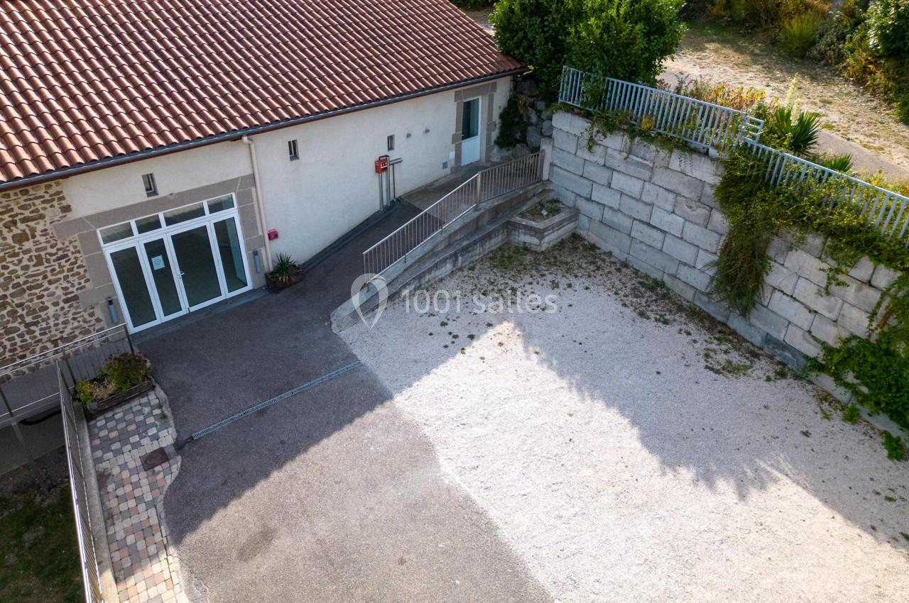 Entrée d'un bâtiment avec rampe d'accès, espace pavé et gravillonné, entouré d'un mur en pierre et de végétation.