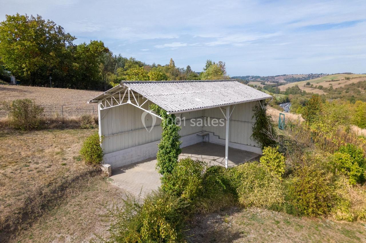 Abri agricole ouvert avec toit en tôle, entouré de végétation, situé dans un paysage rural vallonné.