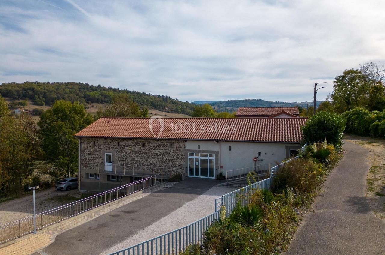 Bâtiment en pierre avec toit en tuiles rouges, rampe d'accès et végétation environnante, sous un ciel partiellement nuageux.
