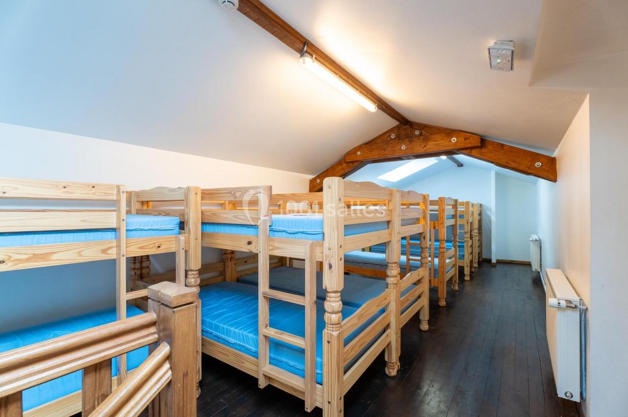 Dortoir avec plusieurs lits superposés en bois, matelas bleus et plafond mansardé avec poutres apparentes.