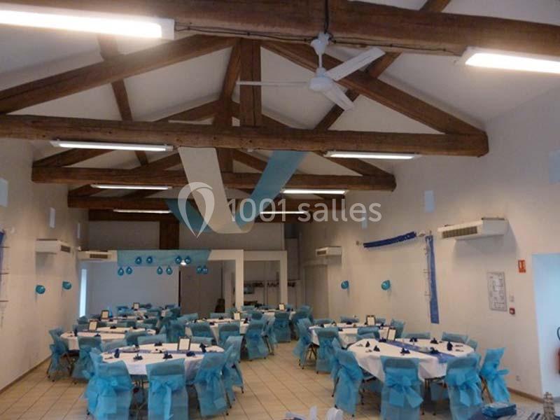 Salle de réception décorée avec des nappes et chaises bleues, poutres apparentes et guirlandes suspendues.