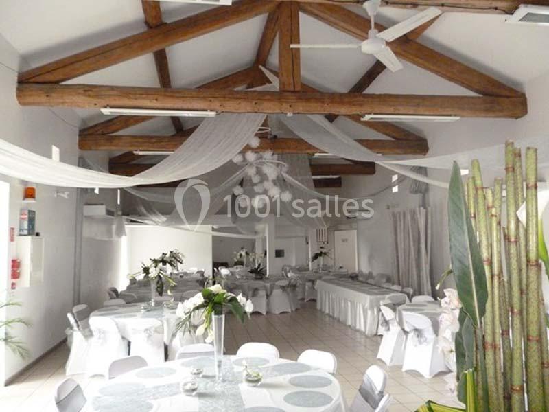 Salle de réception décorée avec des tables rondes et rectangulaires, nappes blanches et poutres apparentes au plafond.
