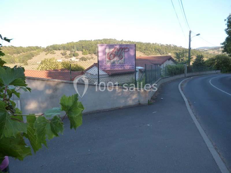 Panneau publicitaire devant un bâtiment entouré de collines, le long d'une route bordée de végétation.