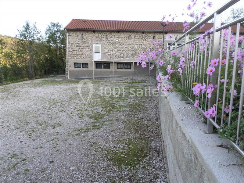 Bâtiment en pierre avec toit en tuiles, entouré d'une cour gravillonnée et de fleurs sur un muret.