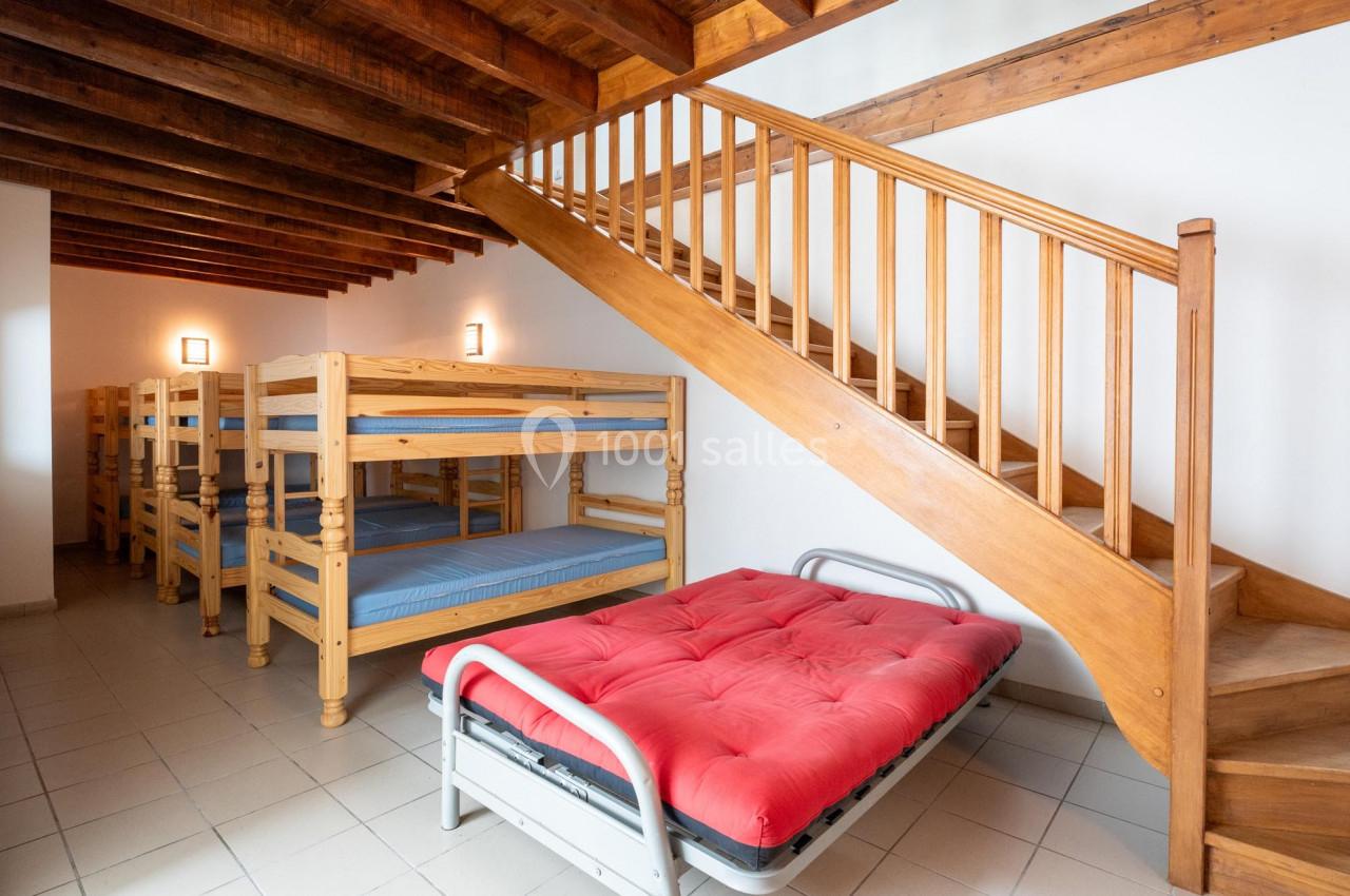 Dortoir avec lits superposés en bois, un lit pliant avec matelas rouge et escalier en bois à droite.