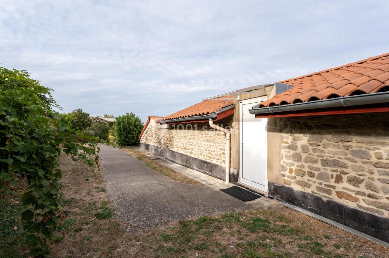 Mur en pierre avec toit en tuiles rouges, chemin asphalté et végétation en arrière-plan sous un ciel partiellement nuageux.