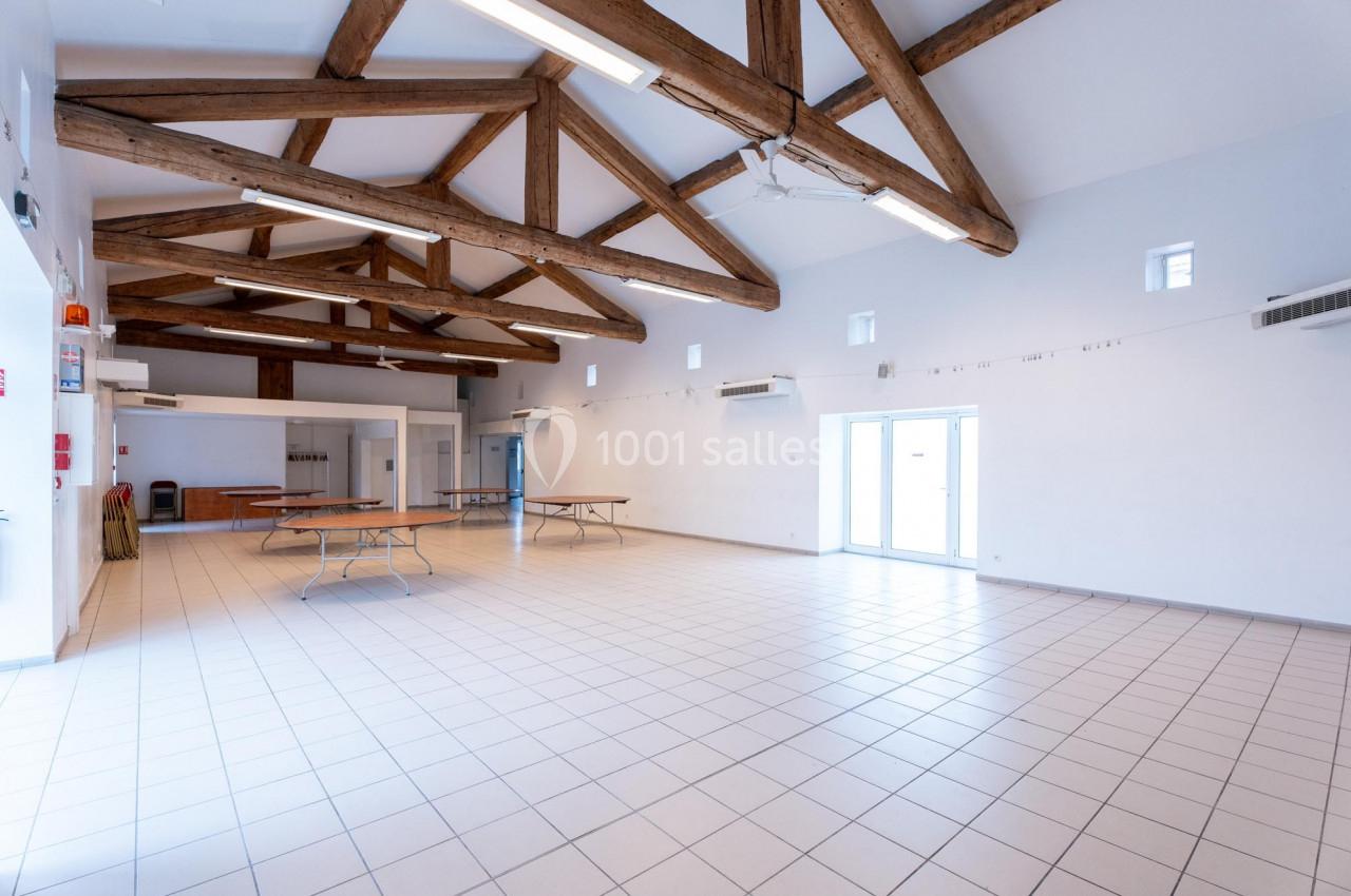 Salle spacieuse avec plafond en poutres apparentes, carrelage clair et tables pliantes contre les murs.