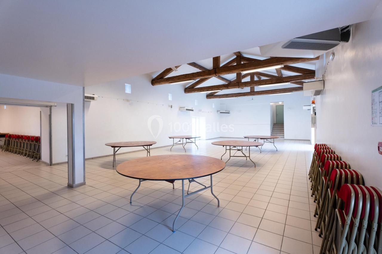 Salle vide avec sol carrelé, tables rondes pliantes espacées et chaises empilées contre les murs, plafond avec poutres…