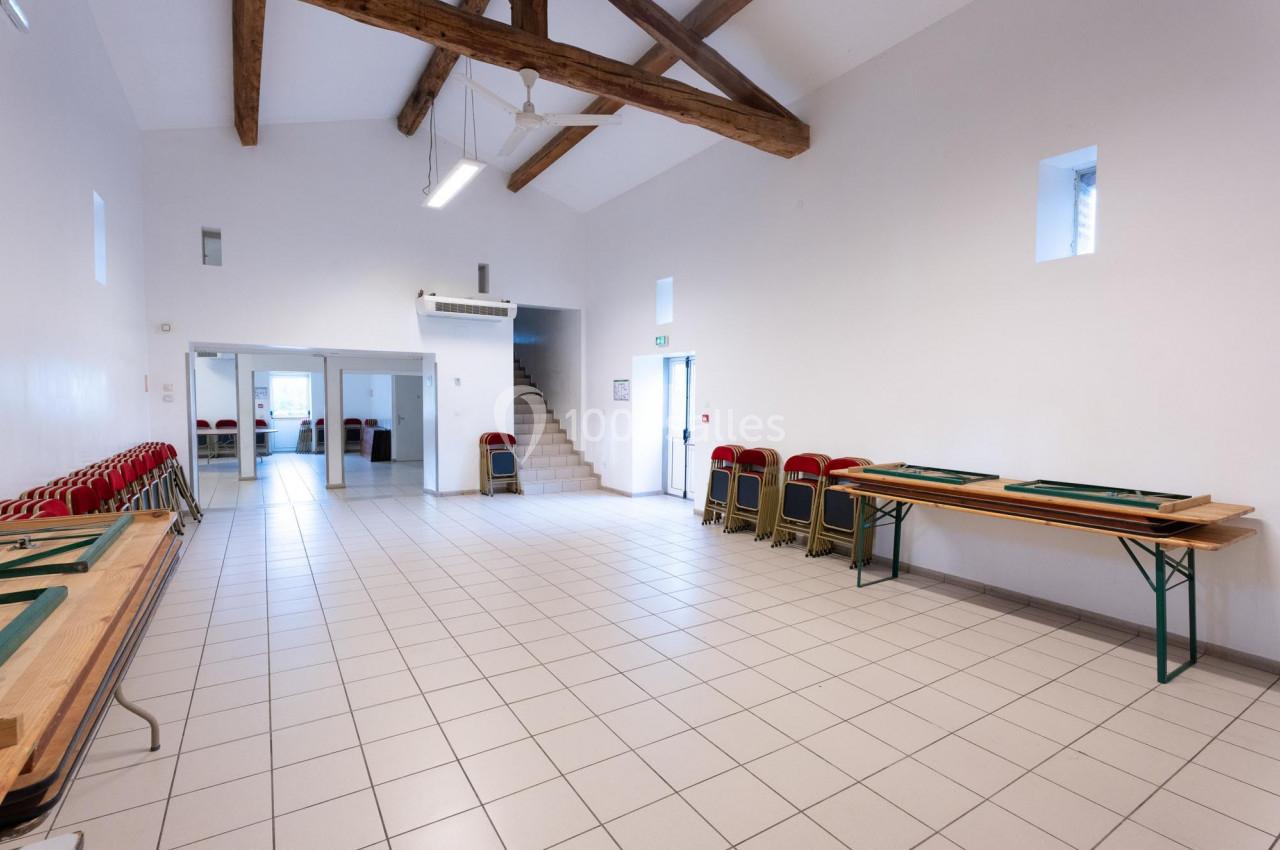 Salle lumineuse avec carrelage clair, poutres apparentes, tables pliantes et chaises empilées contre les murs.