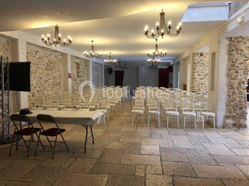 Salle en pierre avec des chaises blanches alignées, une table à l'avant et des lustres suspendus au plafond.