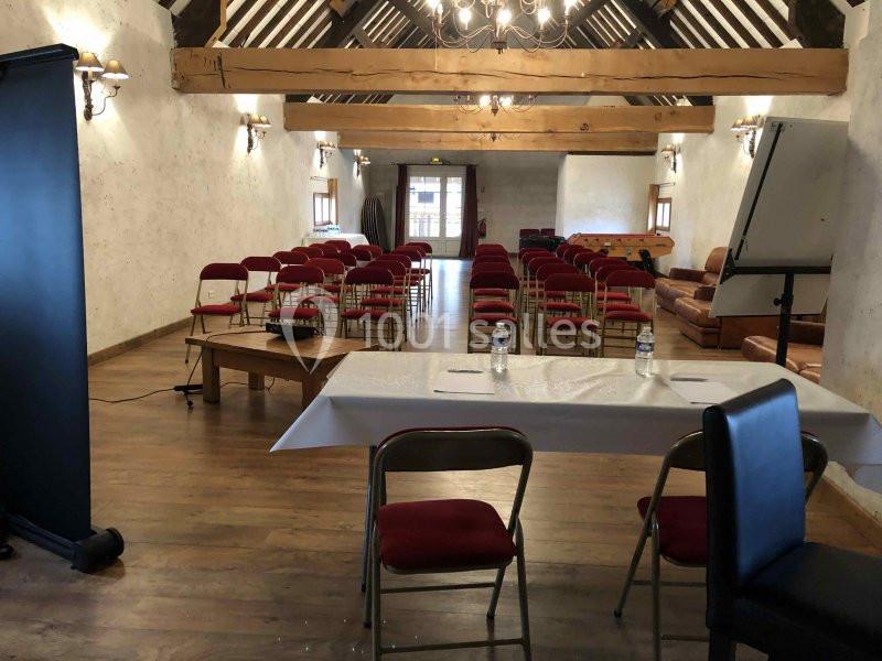 Salle de réunion avec des rangées de chaises rouges, une table blanche à l'avant et un plafond en poutres apparentes.