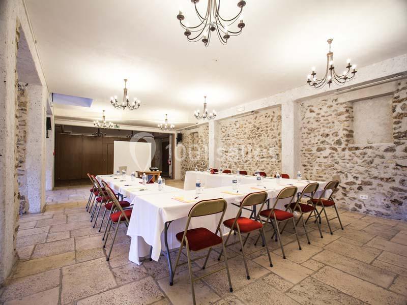 Salle de réunion avec murs en pierre, tables disposées en U, chaises rouges et éclairage par lustres.