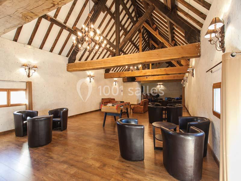 Salle spacieuse avec poutres apparentes, parquet en bois, fauteuils en cuir, baby-foot et éclairage chaleureux.