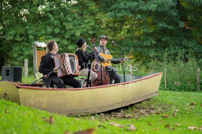 Trois musiciens jouent de l'accordéon, de la contrebasse et de la guitare dans une barque posée sur l'herbe.