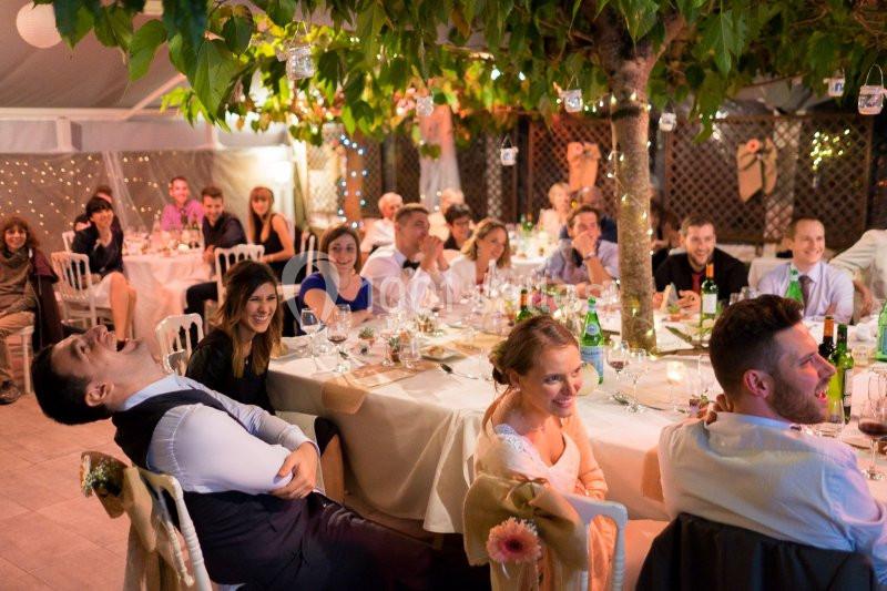 Des invités assis autour de tables décorées, riant et écoutant une intervention lors d'une réception festive en soirée.