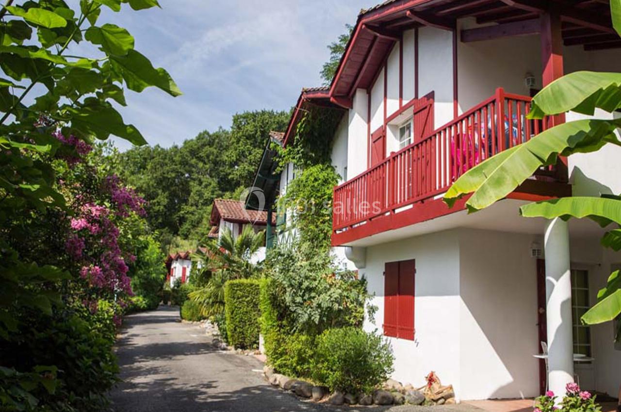 Maison basque traditionnelle avec volets rouges, balcon en bois et jardin verdoyant, bordant une allée ombragée.
