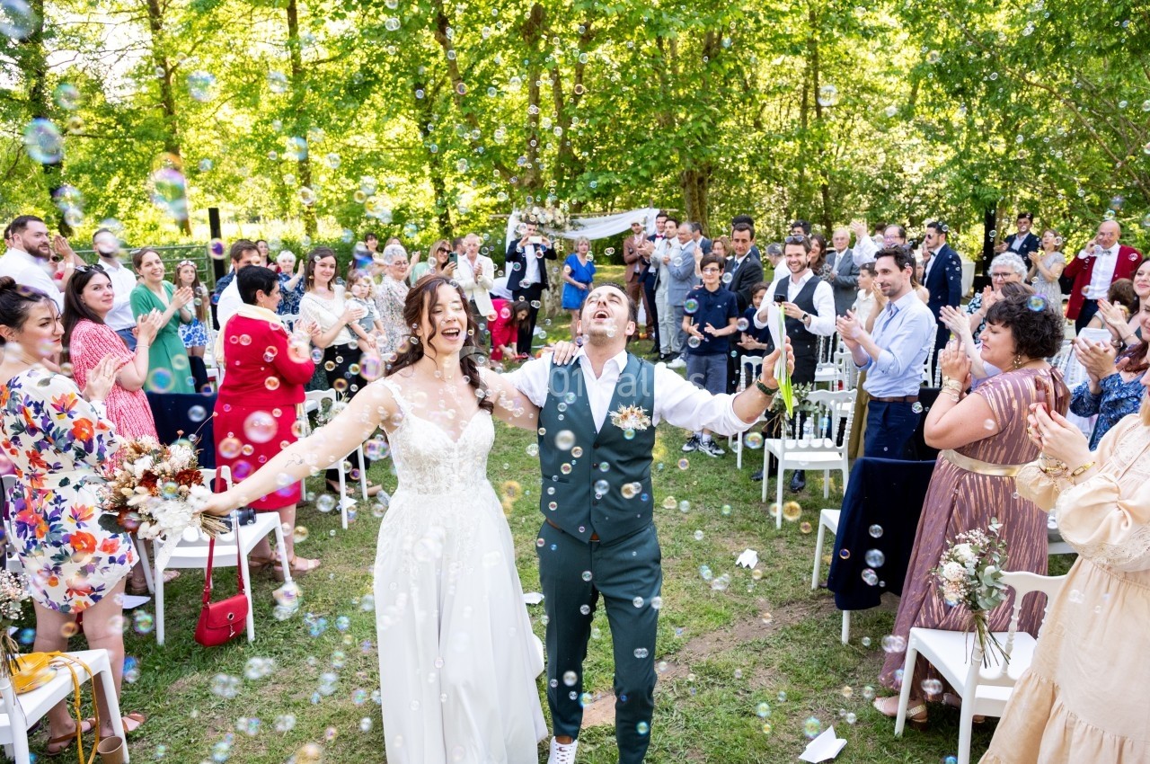 Un couple de mariés souriant entouré d'invités applaudissant dans un jardin, avec des bulles flottant dans l'air.