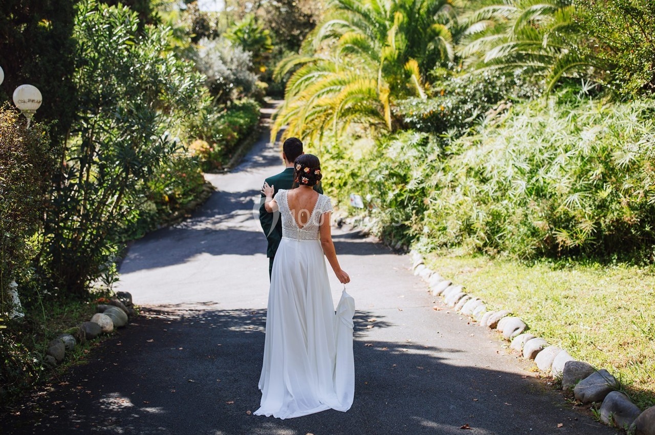 Une mariée en robe blanche marche main dans la main avec un homme en costume sur une allée bordée de végétation luxuriante.