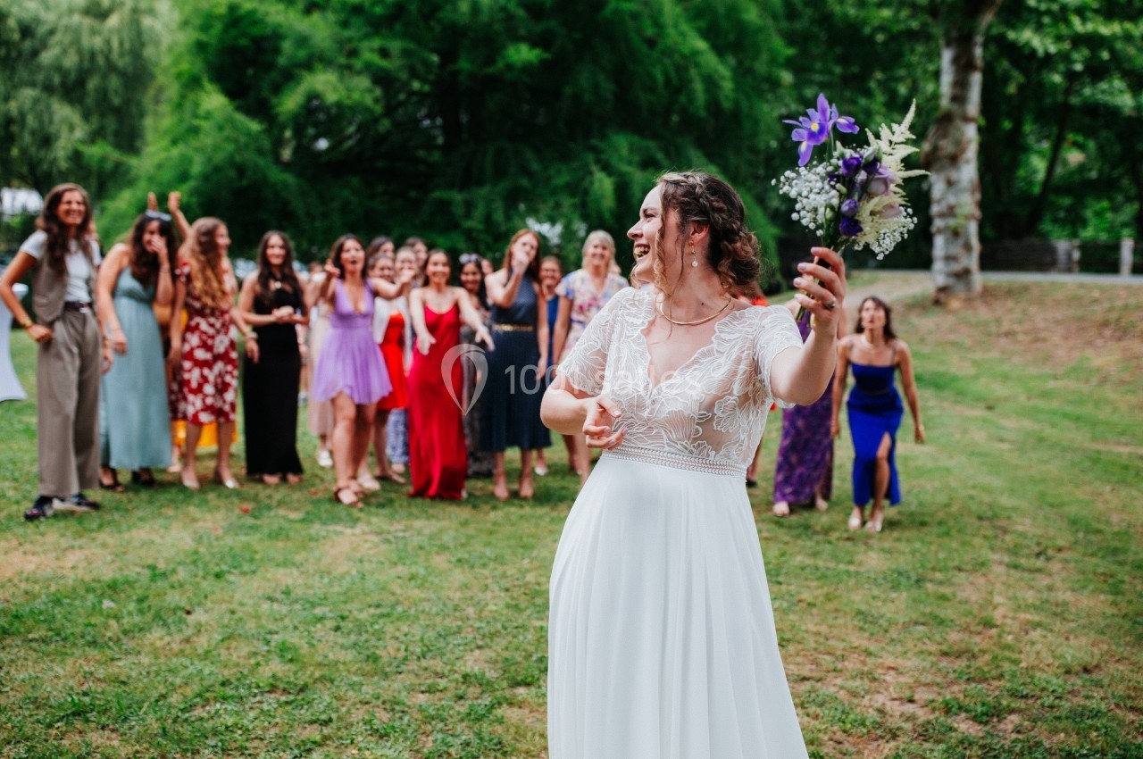 Une mariée en robe blanche lance son bouquet à un groupe de femmes rassemblées sur une pelouse.