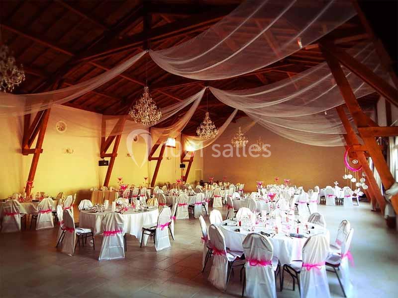 Salle de réception décorée avec des tables rondes, nappes blanches, chaises ornées de rubans roses et plafonds drapés.