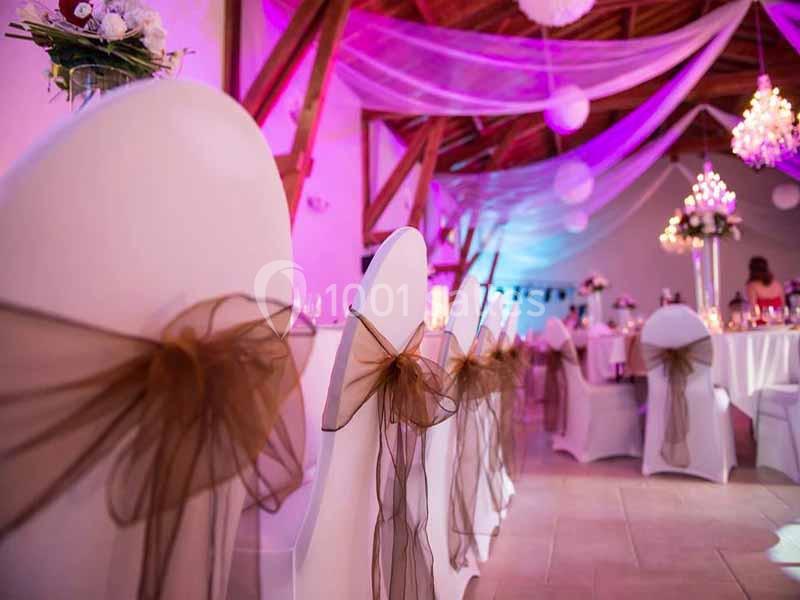 Chaises blanches décorées de nœuds en organza brun dans une salle de réception illuminée de lumières roses et violettes.