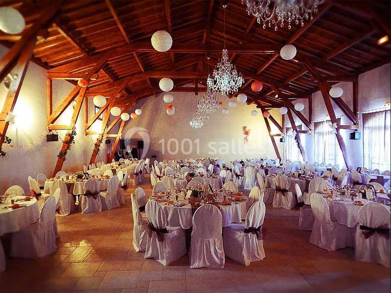 Salle de réception décorée avec des tables rondes dressées, chaises blanches et lanternes suspendues au plafond en bois.