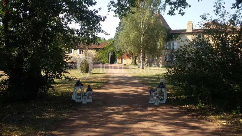 Allée en terre bordée d'arbres menant à des bâtiments anciens avec des lanternes décoratives au premier plan.