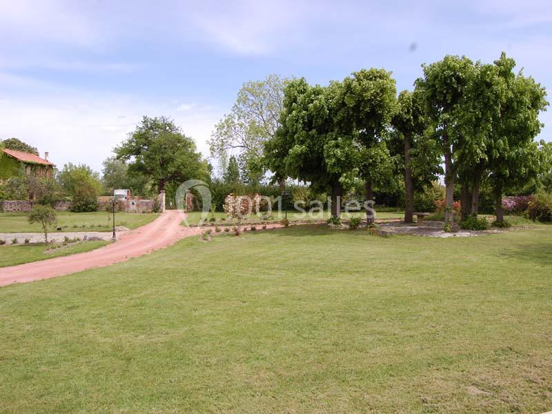 Allée en terre bordée de pelouses et d'arbres, menant à une maison entourée de verdure sous un ciel dégagé.