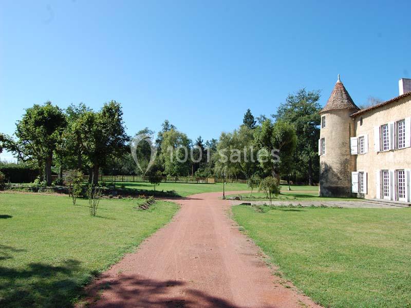 Allée en gravier rouge menant à un bâtiment ancien avec tour, entouré de pelouses et d'arbres sous un ciel bleu.