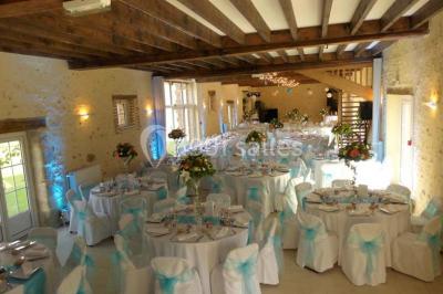 Salle de réception lumineuse avec tables rondes décorées, chaises blanches et poutres apparentes en bois.