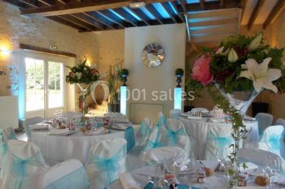 Salle de réception lumineuse avec tables rondes décorées, chaises blanches et poutres apparentes en bois.