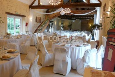 Salle de réception lumineuse avec tables rondes décorées, chaises blanches et poutres apparentes en bois.